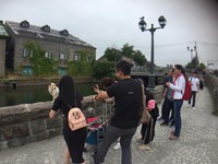 照片中，可成坐在河道旁的矮牆上，面無表情地往左側看，一旁一群遊客一邊激動地揮手吸引注意，一邊舉起相機拍照，畫面相當有趣。
