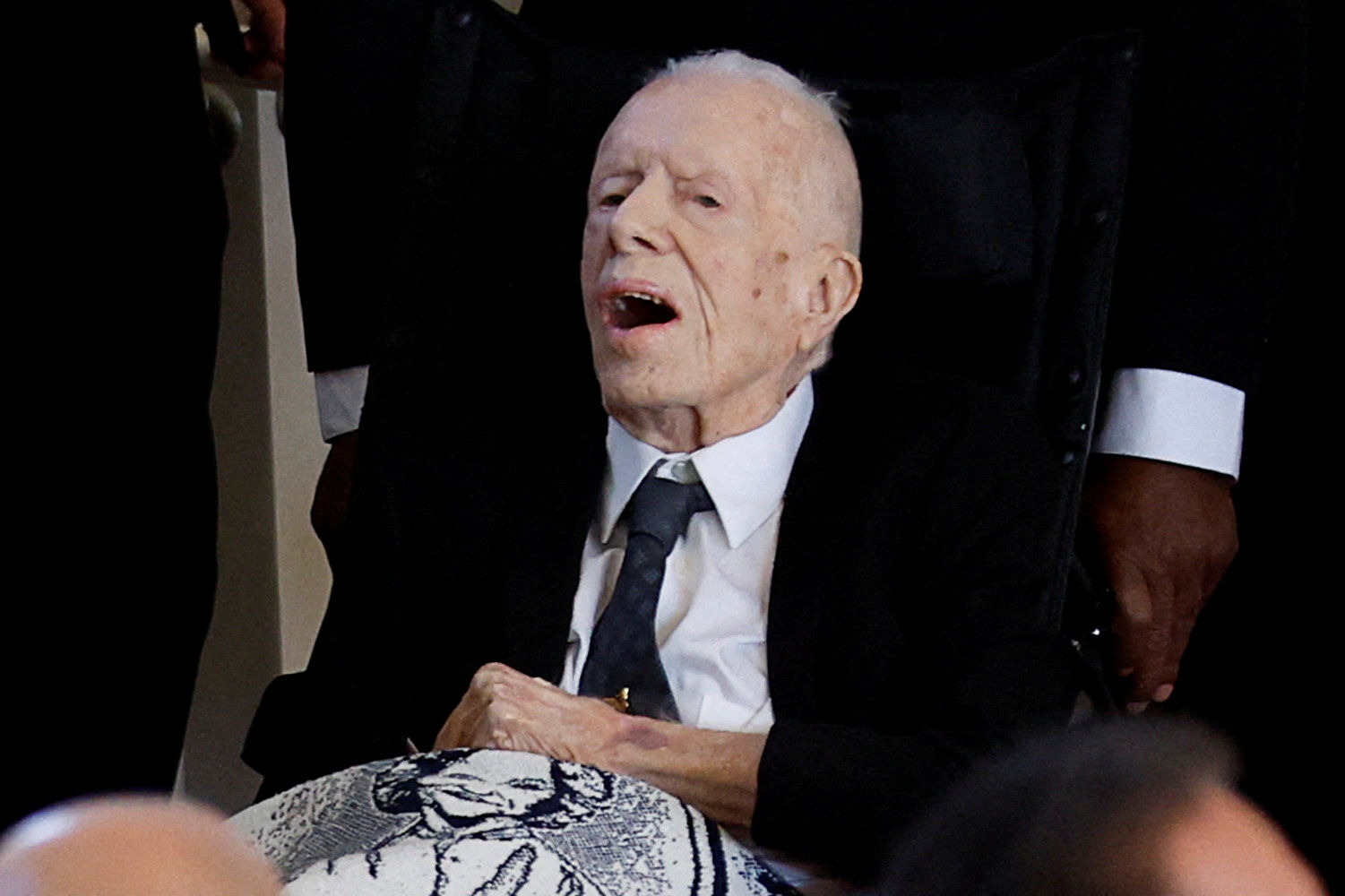 ▲▼美國前總統卡特坐著輪椅、雙腿蓋上印有妻子羅卡琳肖像的毯子，進入哀悼儀式所在的教堂。（圖／路透）